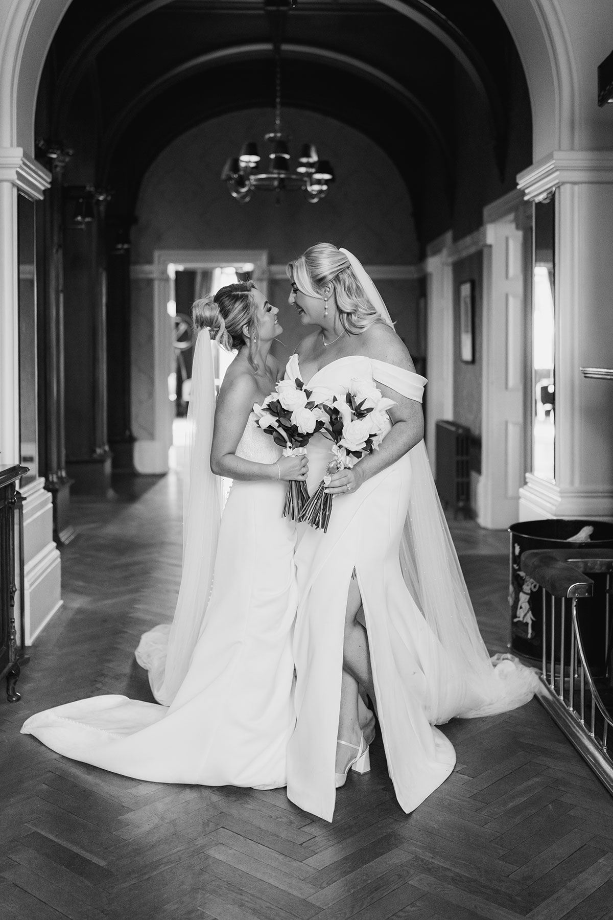 Black and white wedding portrait of two brides holding bouquets and sharing a quiet moment in the venue hallway