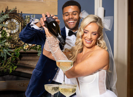 A groom wearing a navy suit jacket and a bride wearing a short wedding dress with tulle gloves and a bow pour prosecco into a tower of glasses