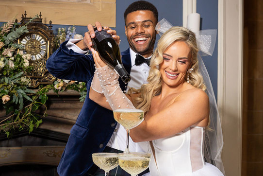 A groom wearing a navy suit jacket and a bride wearing a short wedding dress with tulle gloves and a bow pour prosecco into a tower of glasses