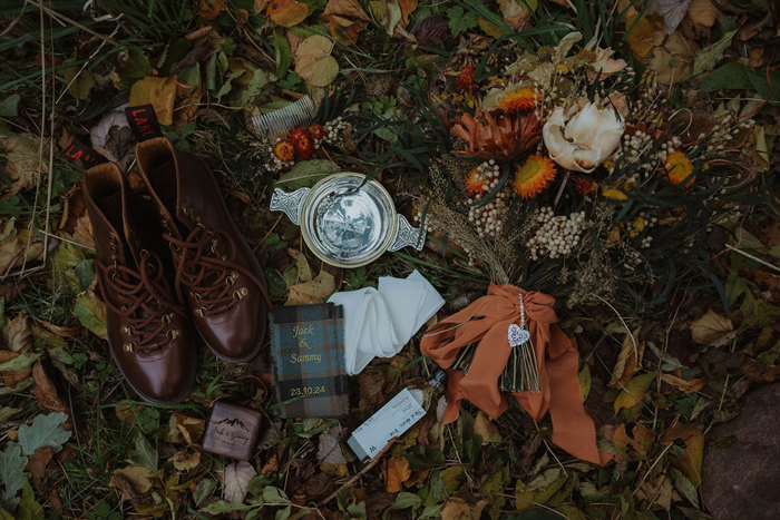 a flat lay of wedding details on leaves including silver engraved quaich, tartan ribbon, wedding rings in box and bouquet of rustic style flowers