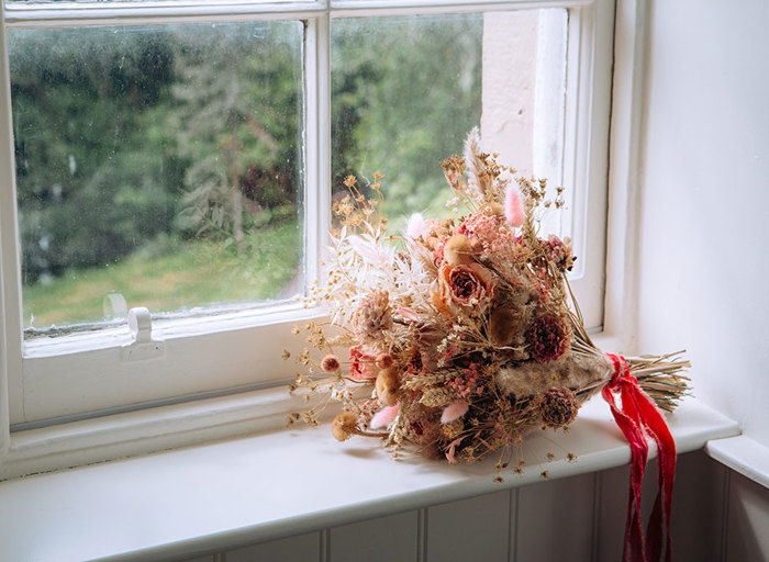 a brown and pale pink dried flower bouquet tied with a bright pink ribbon lies in a windowsill