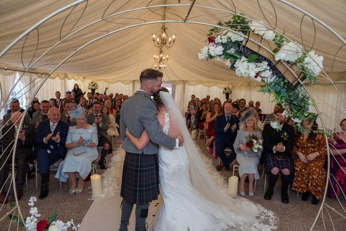 A Bride And Groom Hugging In Front Of A Circular Moongate In A Marquee As Guests Look On