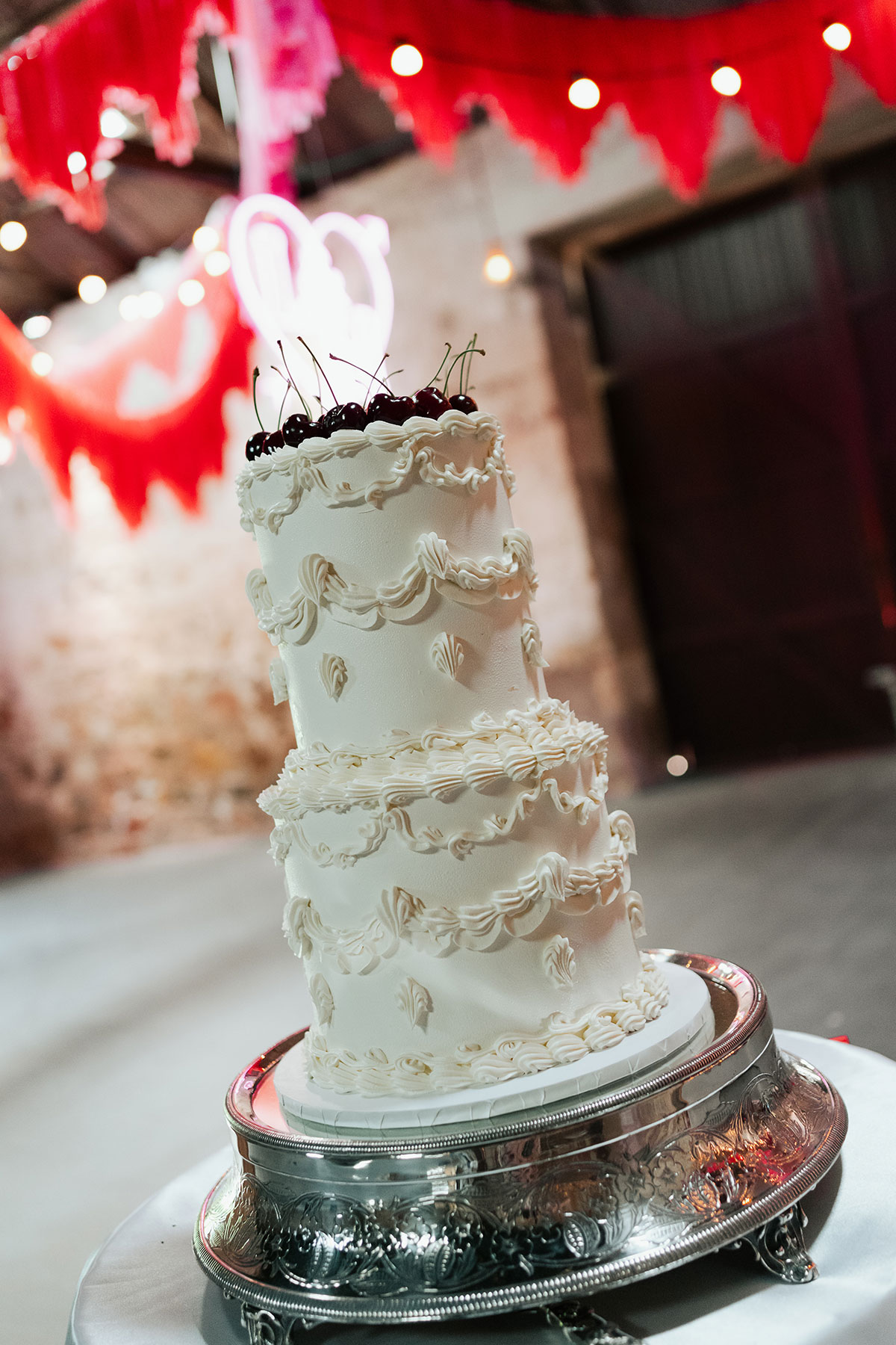 Three-tier vintage-style white wedding cake with piped icing details and cherries on top, displayed at Kinkell Byre beneath red streamers and festoon lighting during a colourful Mexican-inspired wedding in St Andrews.