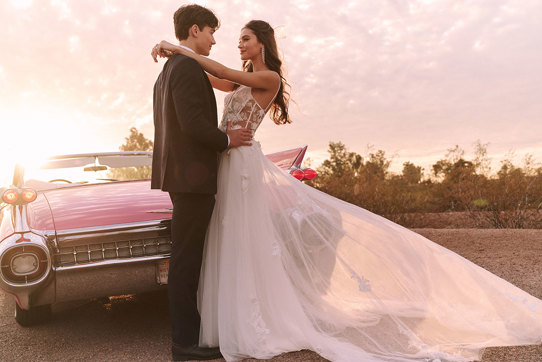 Model wearing corset bodice flowy skirt wedding dress in front of pink corvette with groom next to her