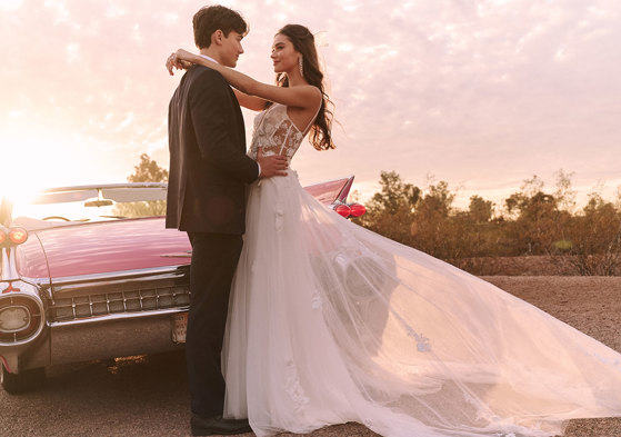 Model wearing corset bodice flowy skirt wedding dress in front of pink corvette with groom next to her