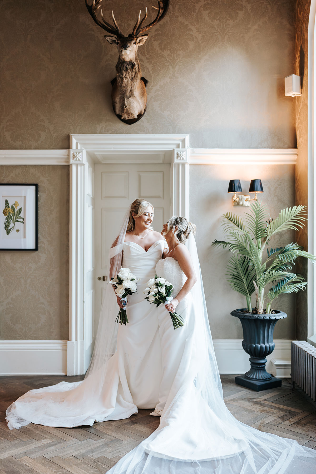 Two brides sharing a smile beneath a mounted stag's head during portraits inside their Scottish wedding venue