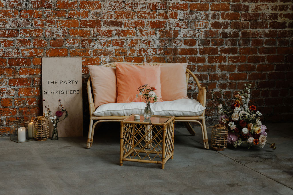 a wicker love seat with coral and pastel pink cushions is surrounded by small wildflower floral arrangements and a sign that says 'The party starts here'