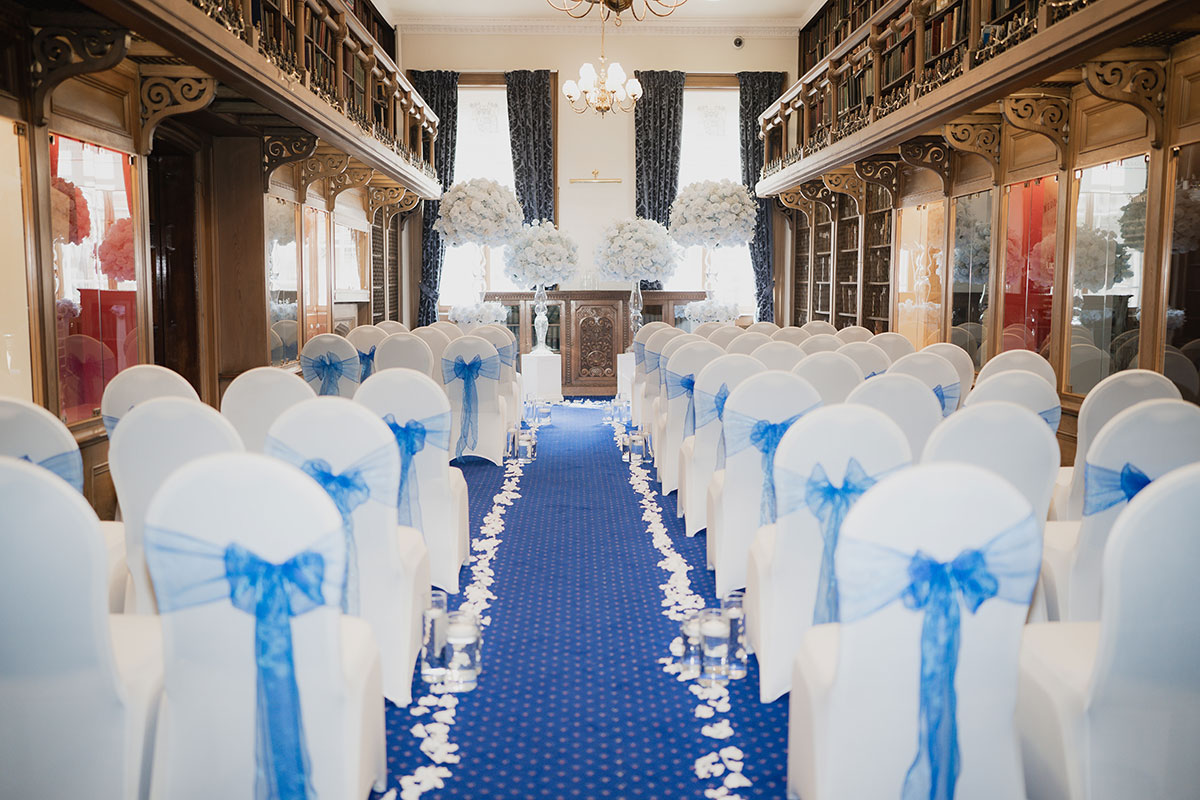 Wedding ceremony setup in the historic library at 1599 at the Royal College featuring a blue aisle, white chair covers with blue bows, floral arrangements and candlelit aisle décor