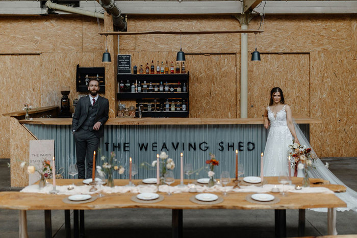 bride and groom stand at either ends of 'The Weaving Shed' labeled bar with a long wooden dining table all set up in front of them