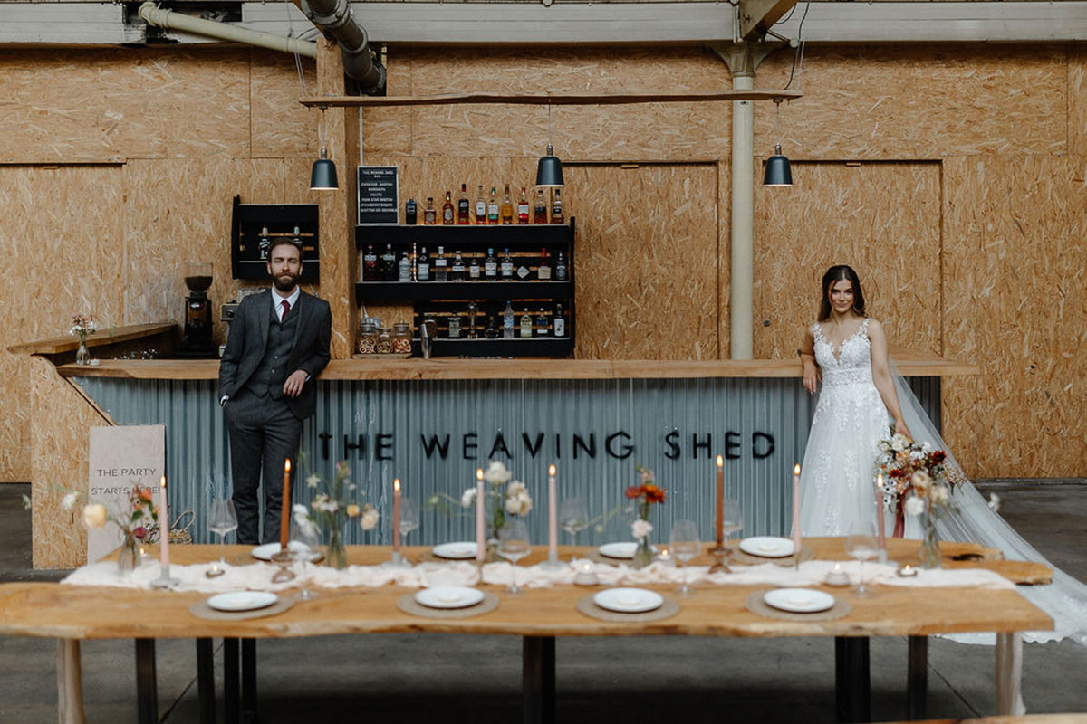 bride and groom stand at either ends of 'The Weaving Shed' labeled bar with a long wooden dining table all set up in front of them