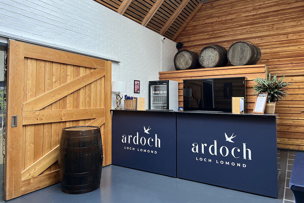 a large wooden barn door and a wooden barrel are placed next to a dark blue bar counter with the name 'Ardoch Loch Lomond' written on the side