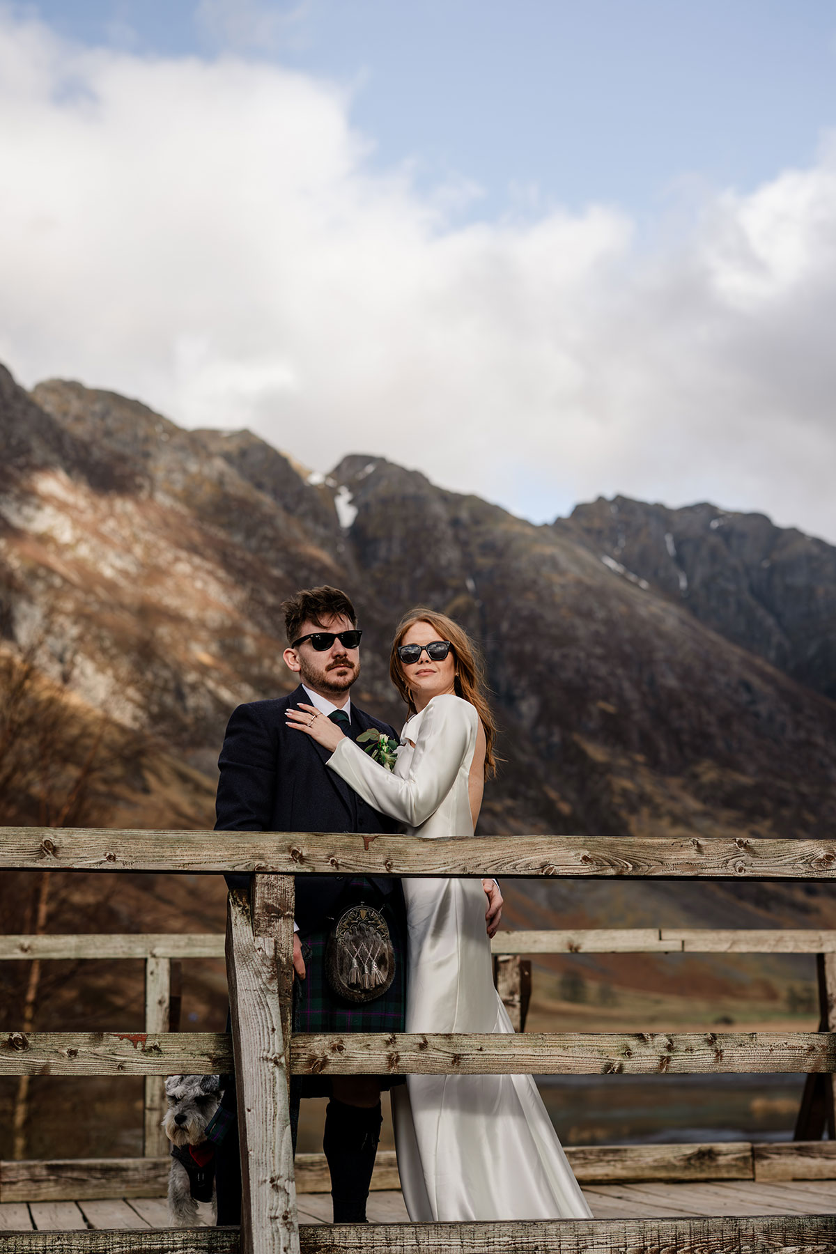 Bride and groom wearing sunglasses on a wooden bridge in Glencoe, with their schnauzer dog and mountain scenery after their Scottish elopement