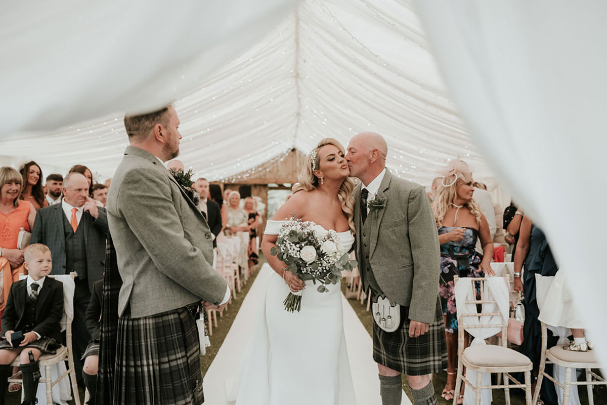 Father kisses the bride on the cheek as she reaches the groom during the marquee ceremony.