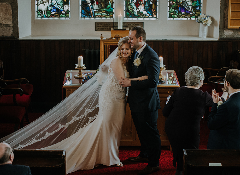 A bride in a fitted dress and long veil and groom in a suit hugging at the alter