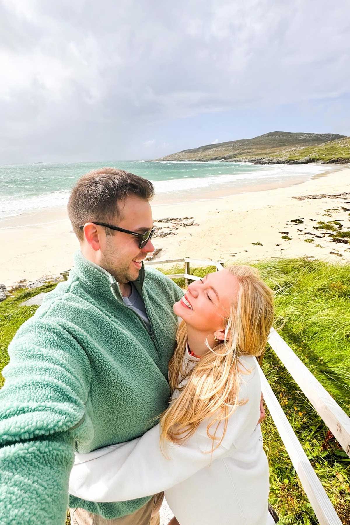 Couple embracing on a windswept beach with turquoise sea and sandy shoreline behind them