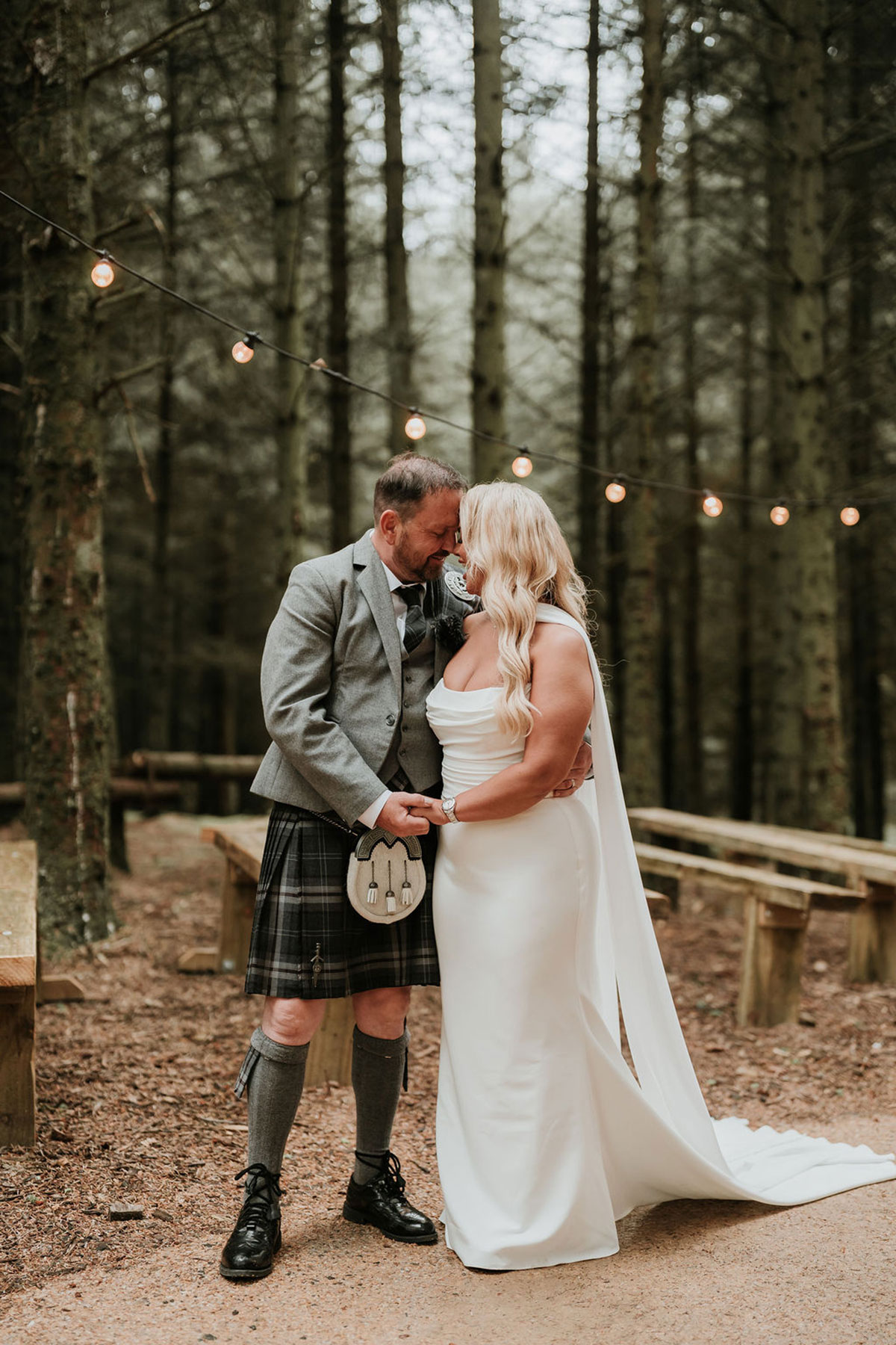 Bride and groom sharing a quiet moment in a woodland clearing with string lights above.