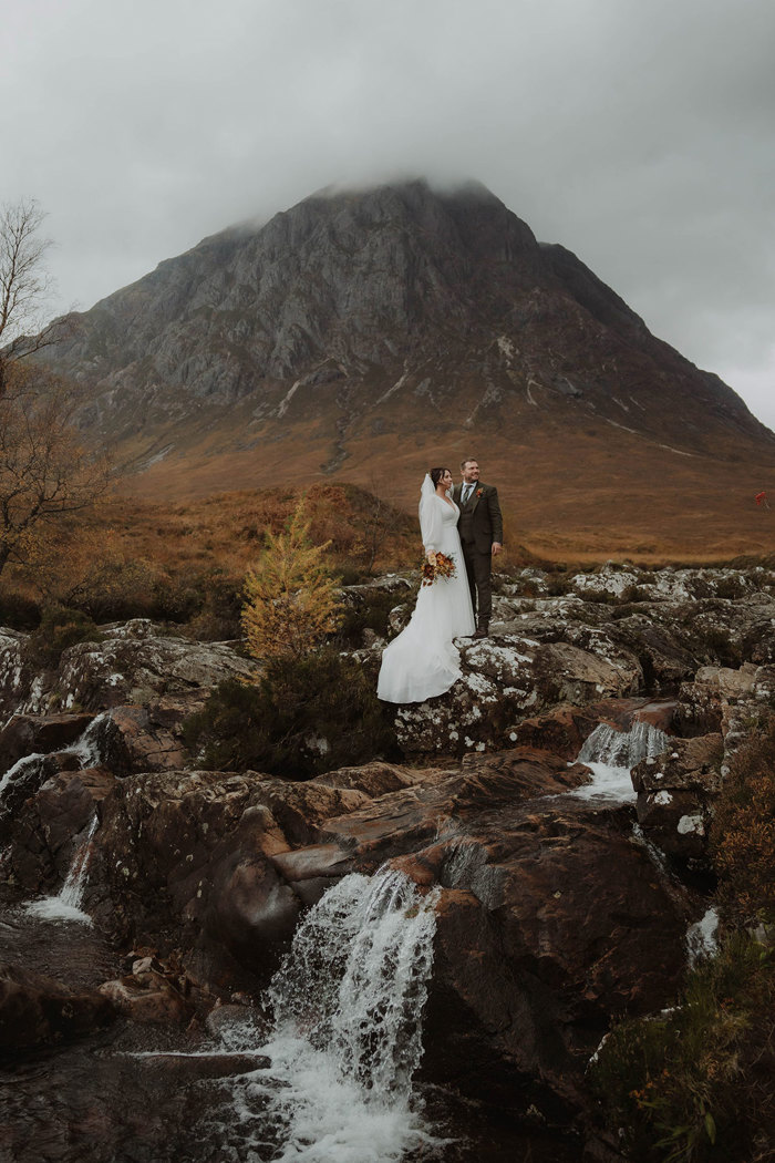 a bride and groom posing by a rocky stream with Buachaille Etive Mòr mountain in the background
