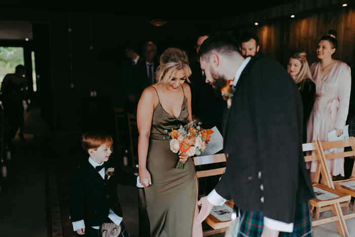 Couple's son holds bridesmaids hand as he walks down the aisle towards his dad