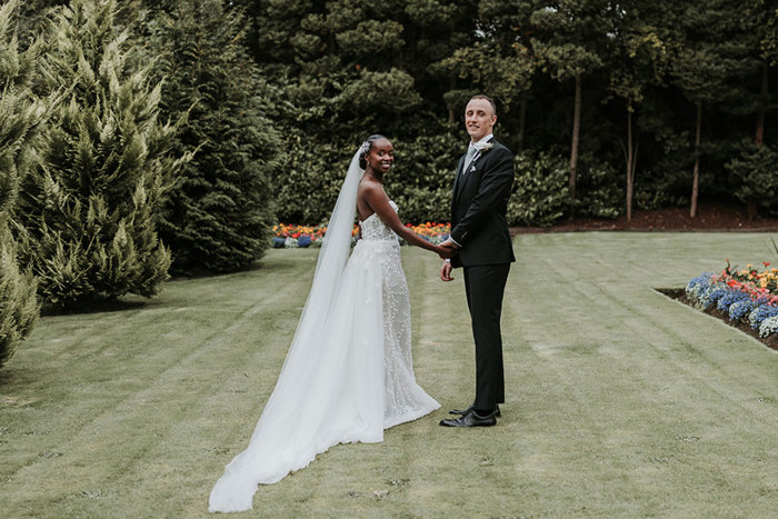 bride in long veil and strapless white dress and groom in black suit stand in garden holding one hand and turning their upper bodies towards the camera