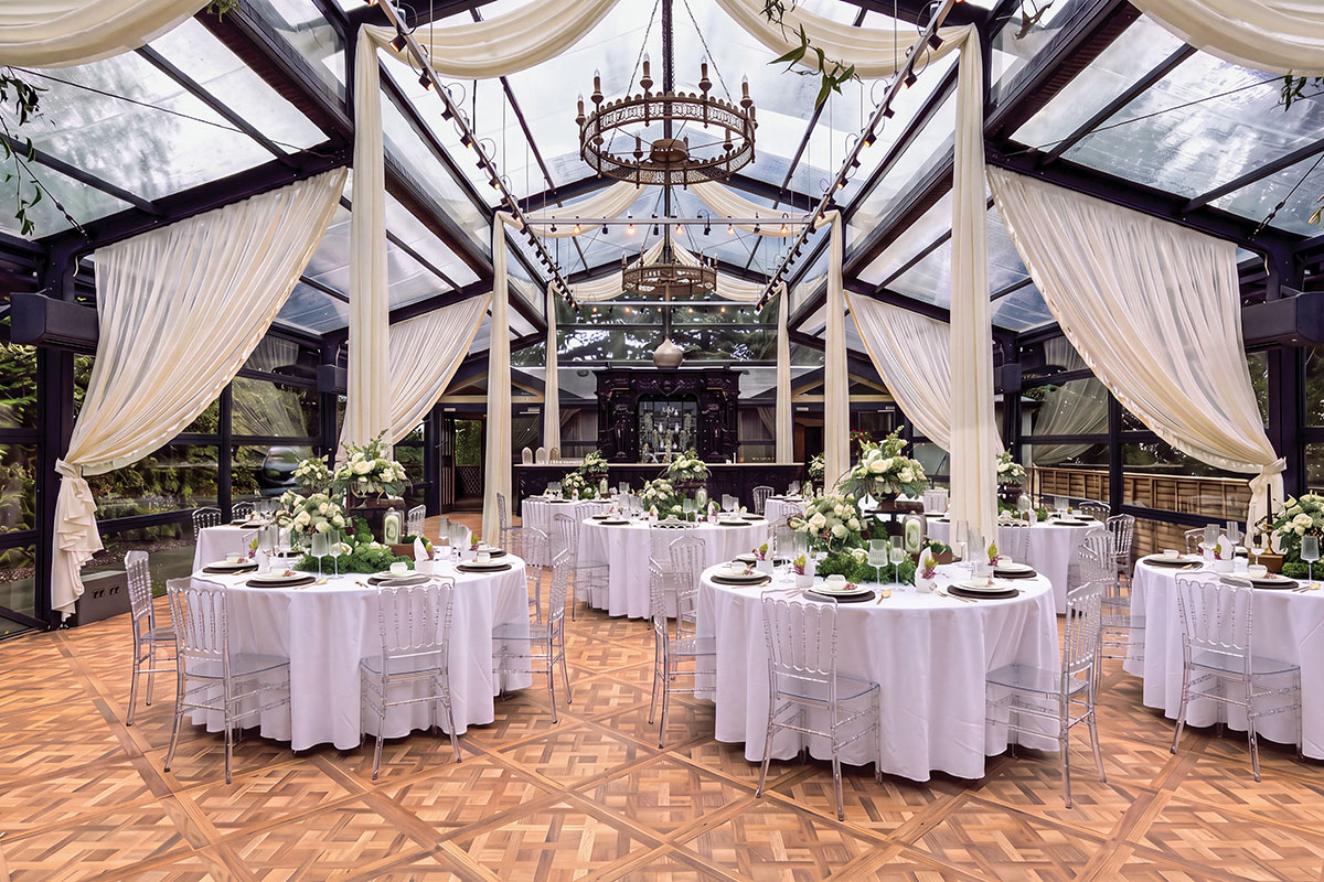 Glass-roofed wedding reception space with white draping, round dining tables, clear chairs and white and green floral arrangements