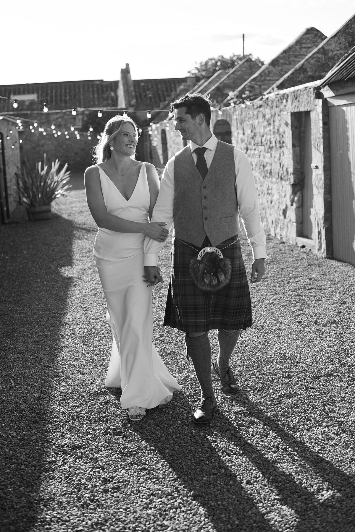 Black-and-white photo of the couple walking hand in hand down a gravel lane at sunset, smiling at each other