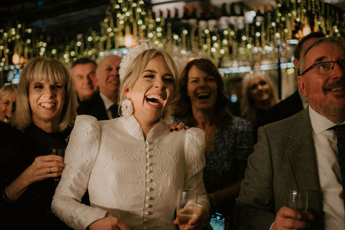 A crowd of people holding champagne flutes and laughing with fairylights behind them