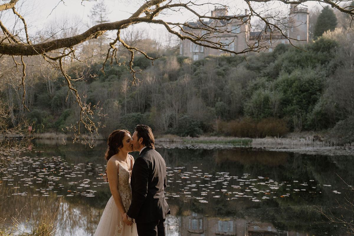 Bride and groom sharing a kiss beside the lake at Auchen Castle, with the castle visible among the trees in the background of the Dumfries and Galloway estate
