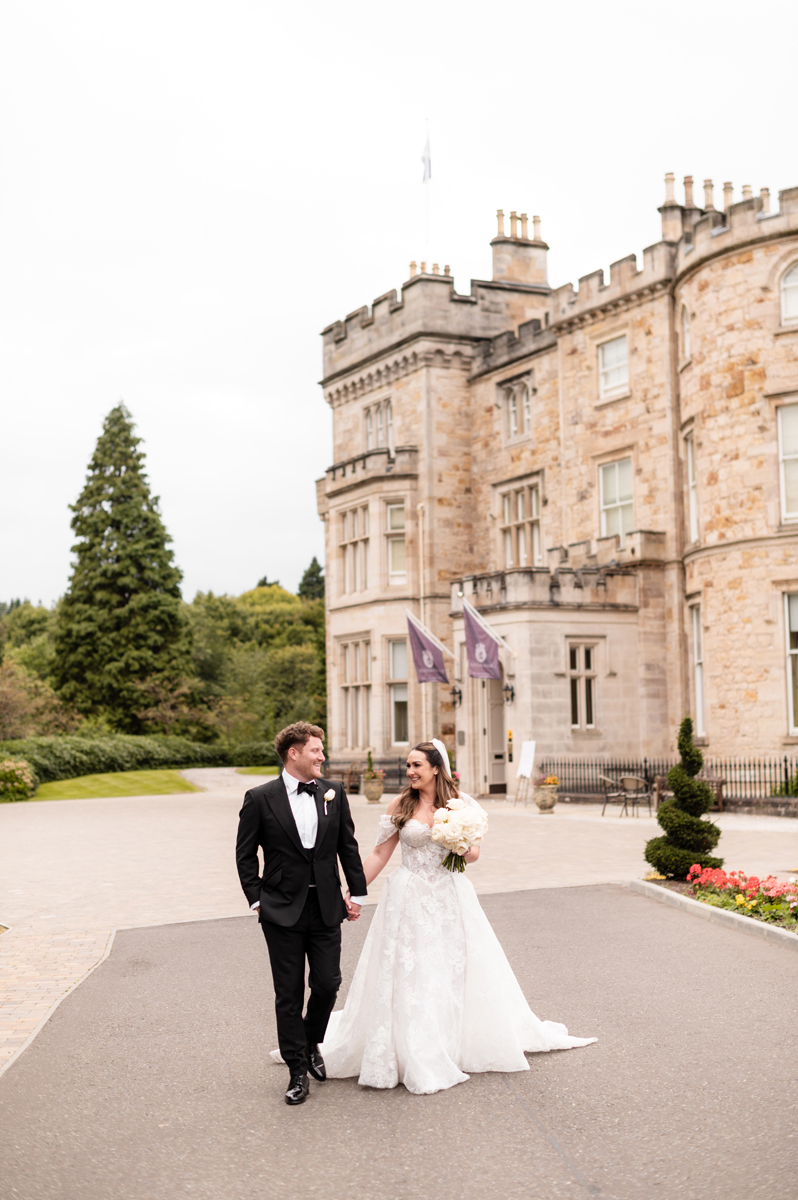 Bride and groom in black-tie wedding outfits walking hand in hand outside Crossbasket Castle near Glasgow.