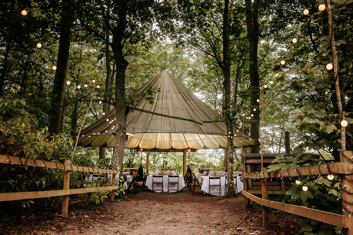 Woodland dining setup at Cambo Estate with long tables beneath a canvas canopy and festoon lighting surrounded by trees