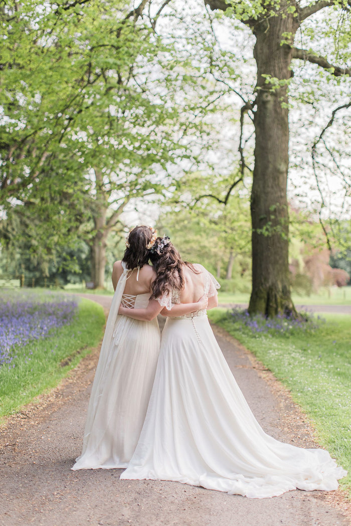 the back of two brides embracing. They are walking on a path with grass and bluebell flowers either side and trees overhead.