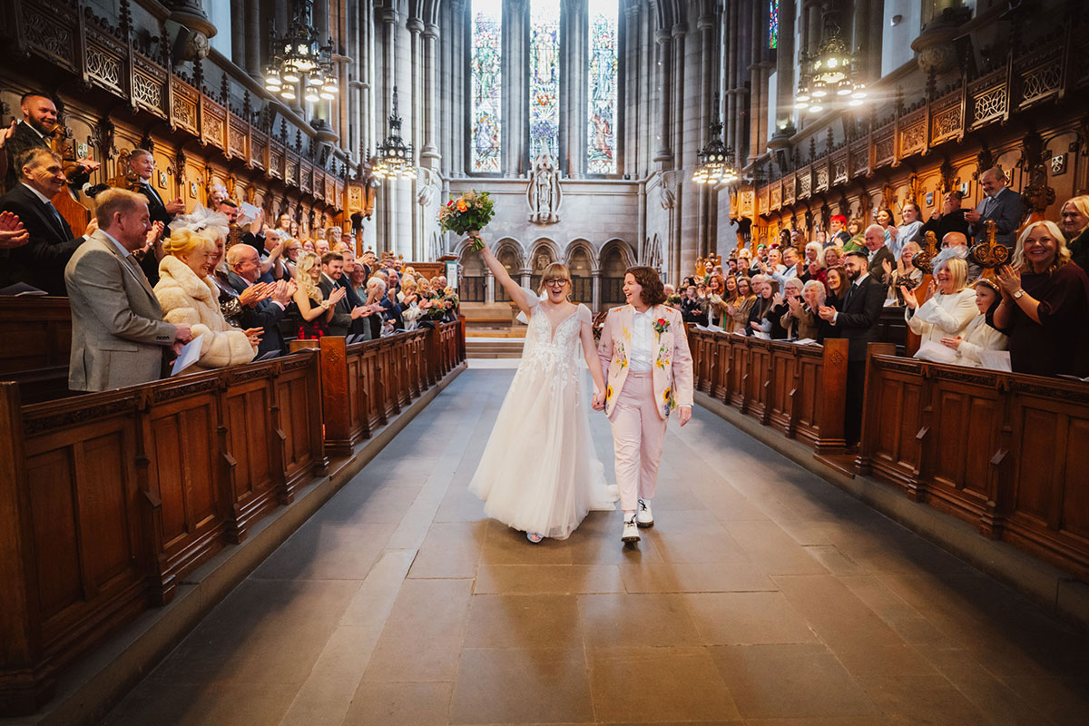 Two brides walking hand in hand down the centre aisle of the University of Glasgow Memorial Chapel as guests applaud, with stained-glass windows and wooden pews surrounding them.