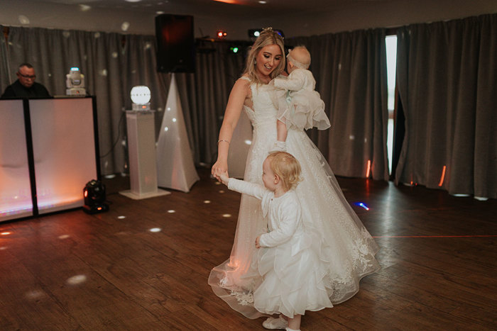 A Bride Dancing With A Baby In Her Arms And A Young Child On The Dancefloor With DJ In Background