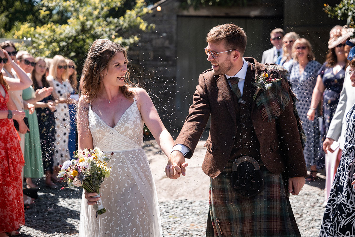 Bride and groom walk through a crowd of guests throwing confetti after the ceremony. The bride wears a lace wedding dress and carries a bouquet of colourful flowers, while the groom wears a brown tweed jacket with tartan kilt and sporran