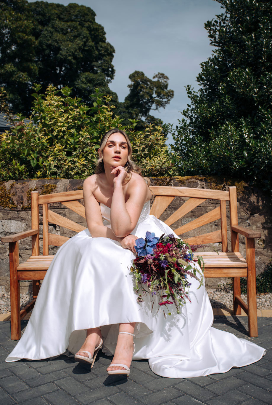 bride in strapless white wedding dress sits on wooden bench holding a bouquet of flowers