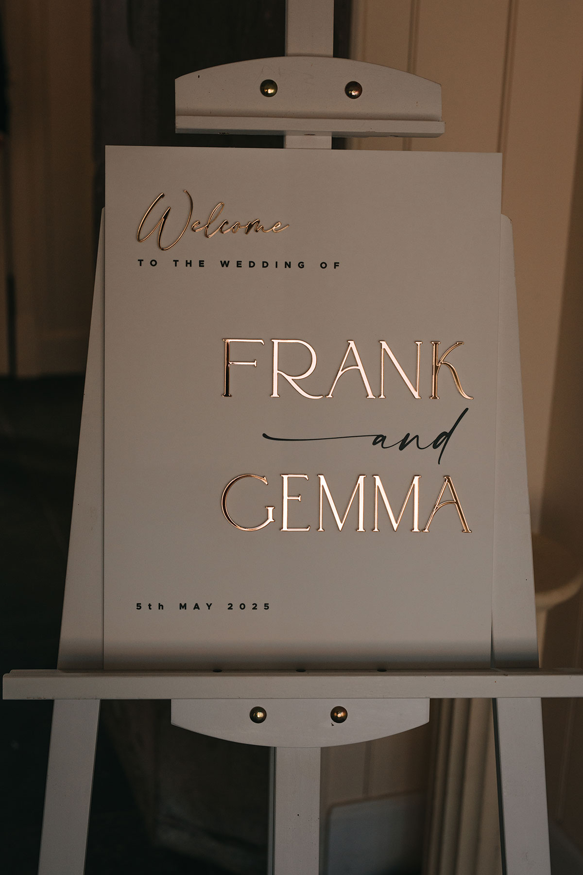 Minimal white wedding welcome sign with gold lettering reading Frank and Gemma on easel