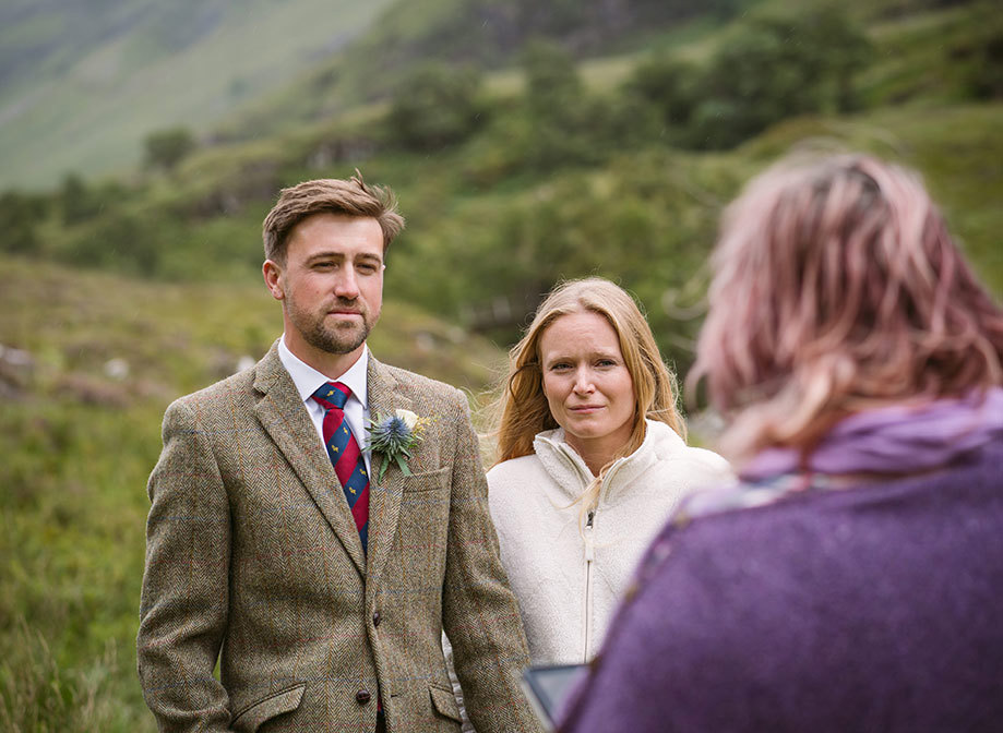 a bride wearing a cream fleece and a groom wearing a green tweed jacket stand in front of a wedding celebrant during a wedding ceremony in Glencoe