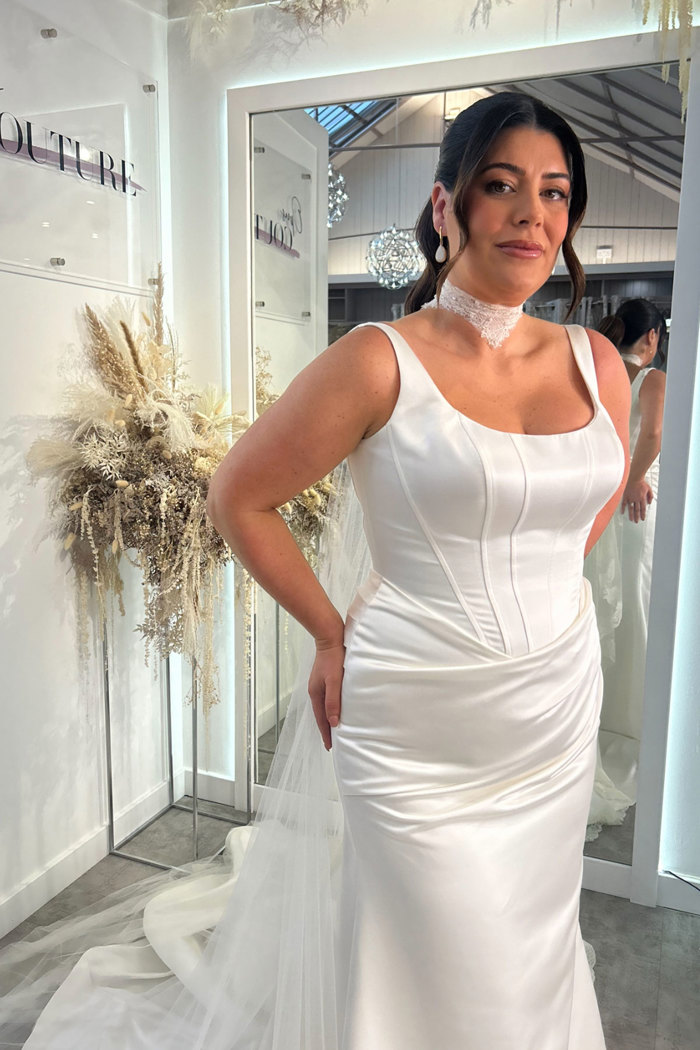 dark-haired bride stands in mirror-filled space, wearing a corset style satin wedding dress and a necktie veil