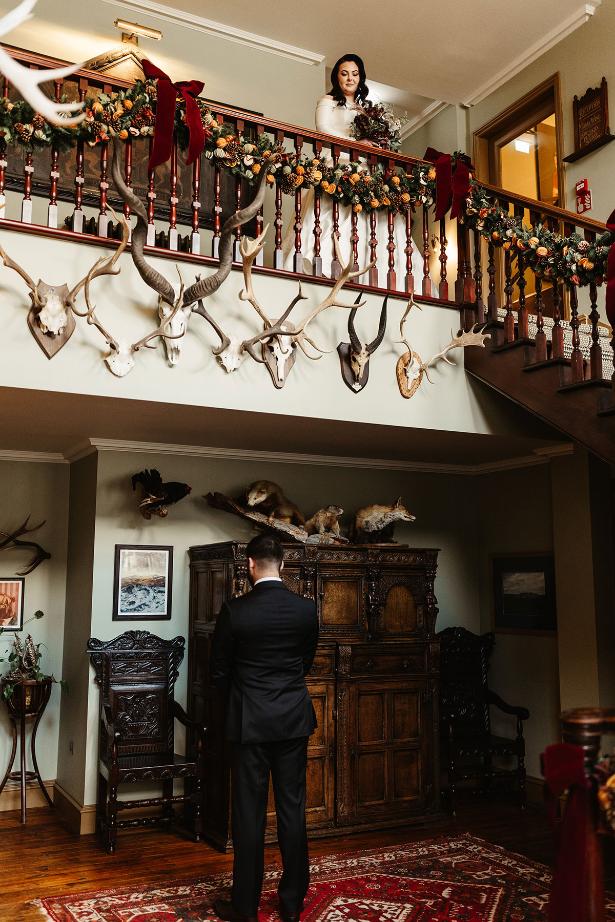 Bride holding her bouquet on a decorated balcony, looking down at the groom waiting below in a taxidermy-filled room with garlands and red velvet bows