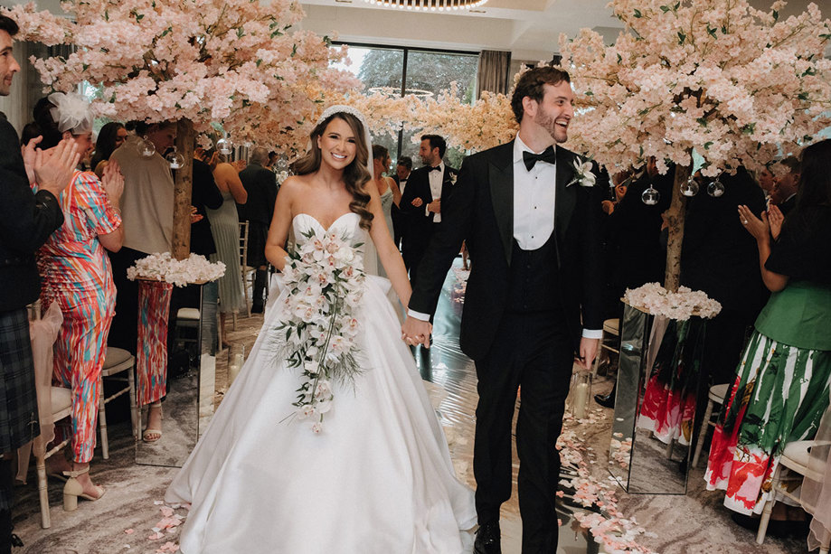 a bride and groom walking up the aisle at Cameron House with cherry blossom trees either side.