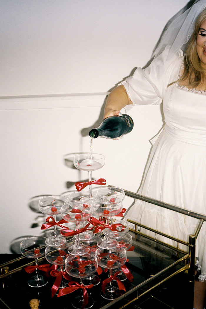 blonde bride pours champagne into champagne tower of coupe glasses with red bows around the stems, all placed on a cocktail trolley