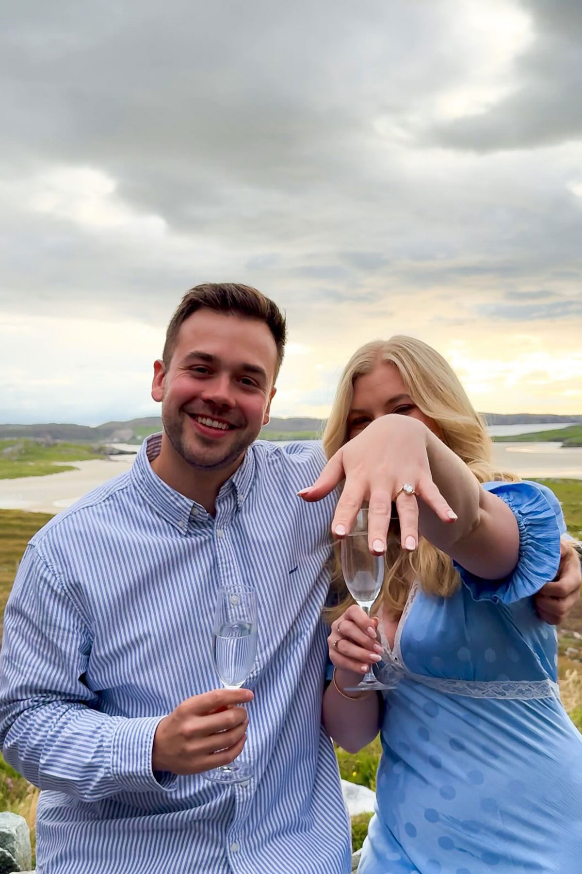 Engaged couple posing with Champagne, woman extending hand to showcase diamond engagement ring against scenic Scottish backdrop