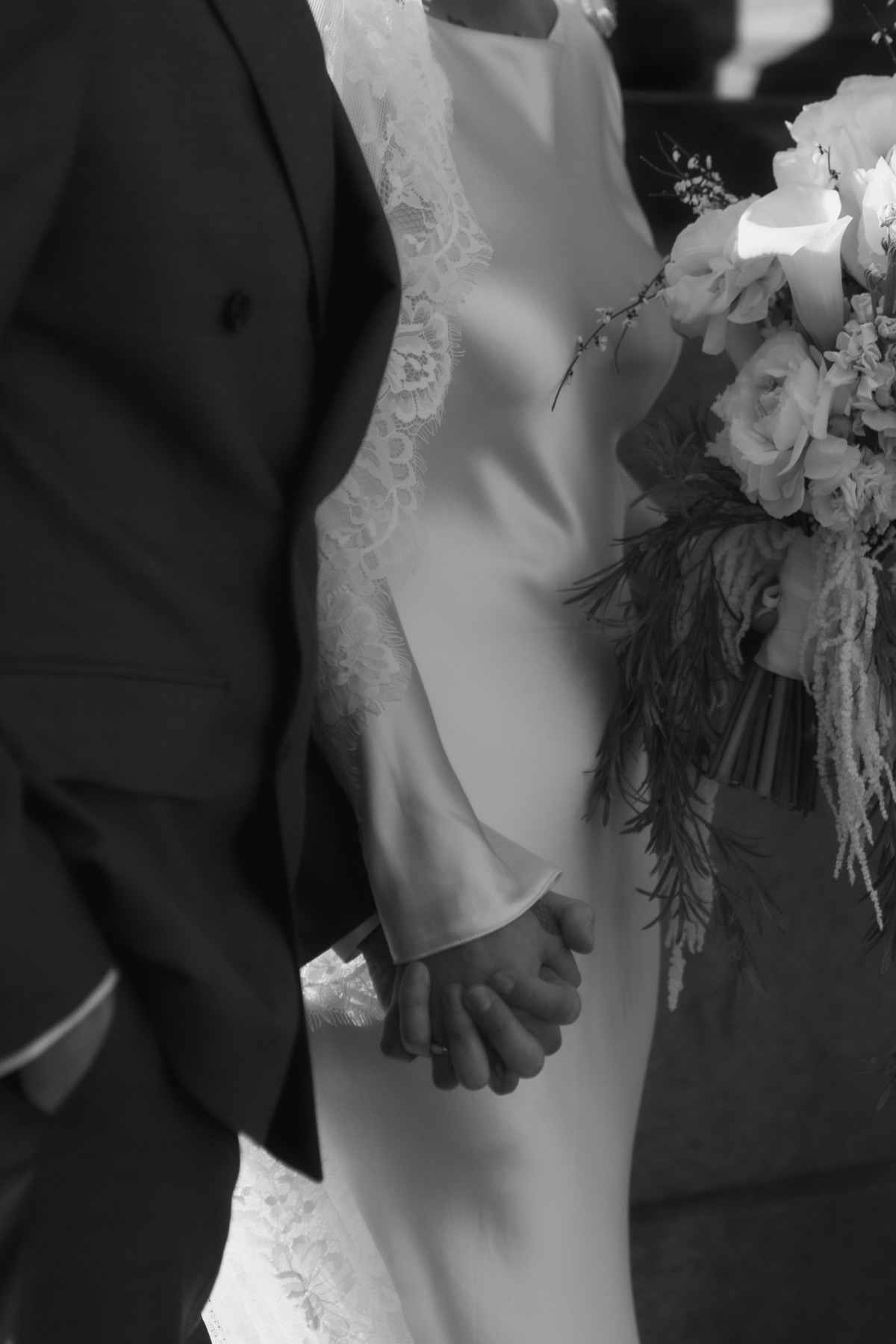 Black-and-white detail of the newlyweds holding hands beside the bride’s white wedding bouquet.