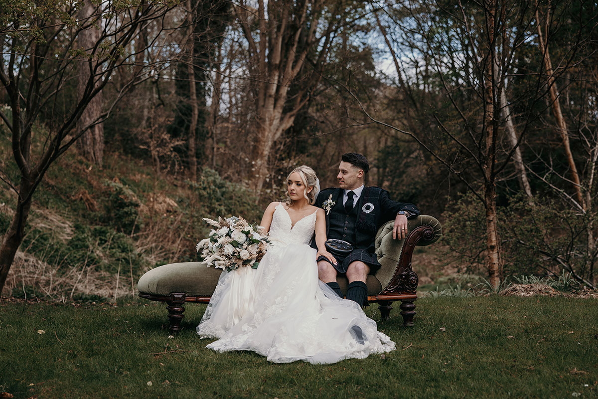 bride in floral embroidered skirt and large neutral bouquet in hand sits next to groom in full kilt outfit on green velvet chaise in a woodland grassy setting
