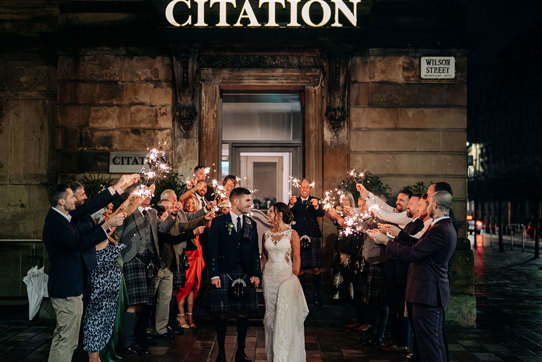 A bride and groom looking at each other as they walk through a crowd of people holding sparklers