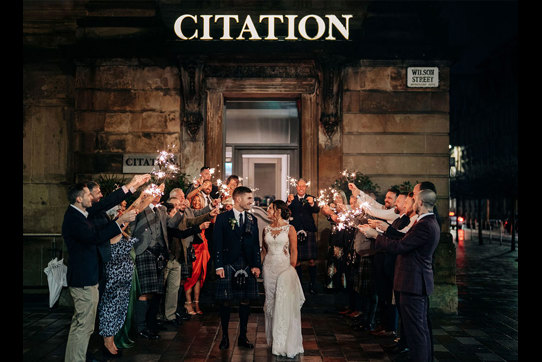 A bride and groom looking at each other as they walk through a crowd of people holding sparklers