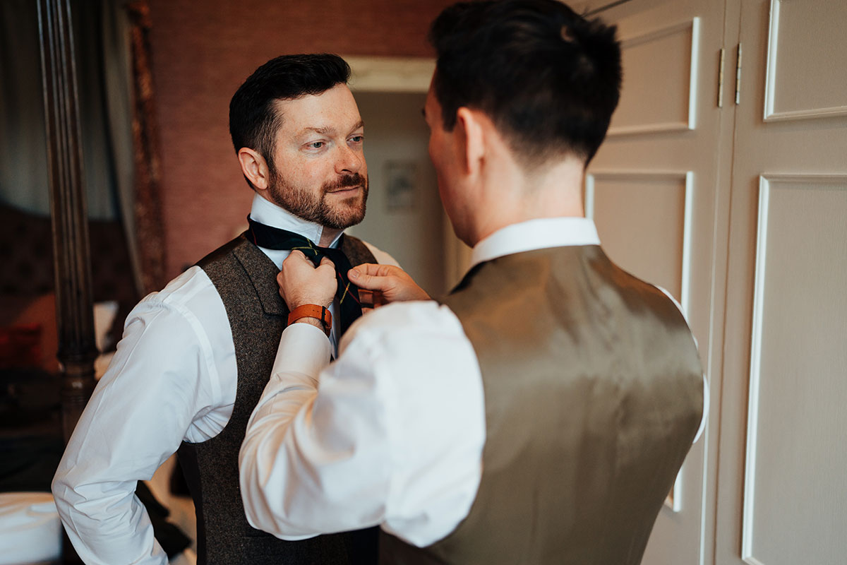 One groom helps the other adjust his tartan tie while getting ready