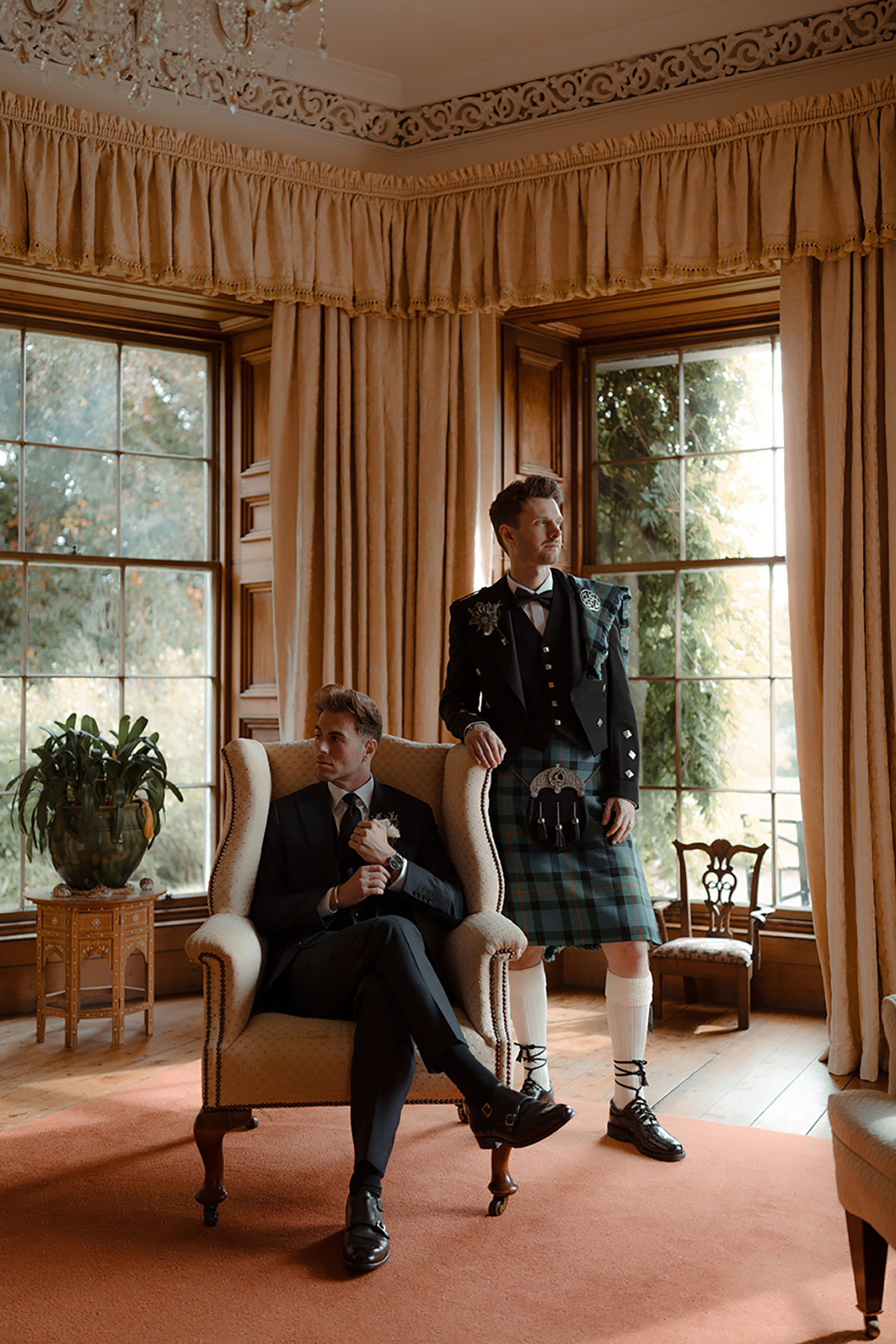 Two grooms pose indoors at Newhall Estate, one seated in a navy suit and the other standing in a Gunn tartan kilt by MacGregor and MacDuff