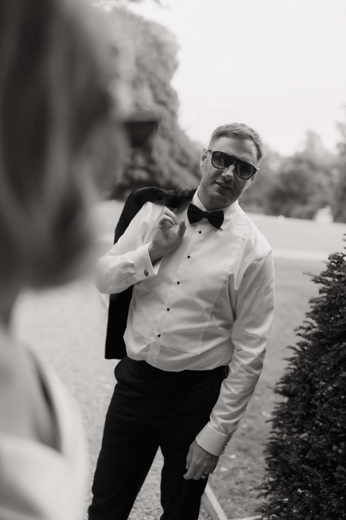 Black-and-white portrait of groom Rory wearing sunglasses and a bow tie in the grounds of Prestonfield House in Edinburgh.