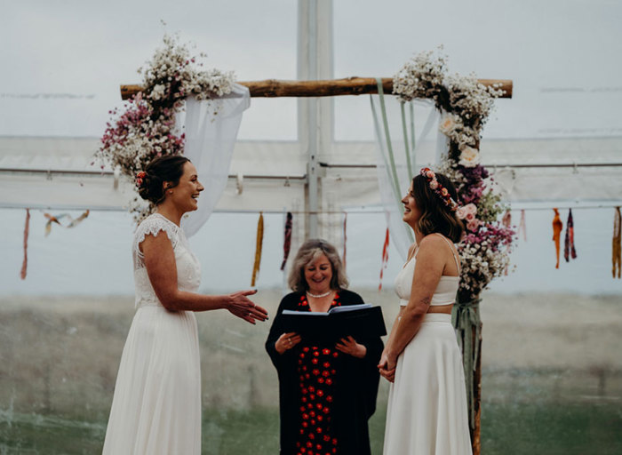 two brides smile as they stand in front of an officiant who is holding a large black folio conducting their wedding ceremony. There is a rectangular wooden frame in the background decorated with and daisy and rose floral arrangement at each corner