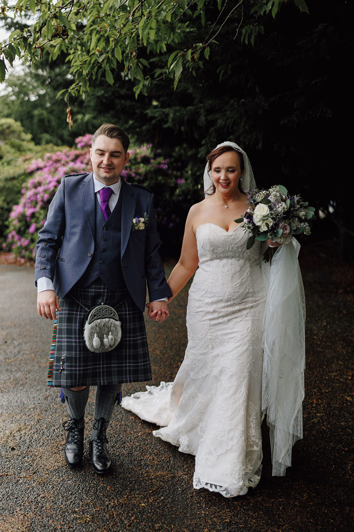 groom in full tartan outfit and bride in strapless floral wedding dress with long veil and bouquet walk hand in hand in front of green trees and bushes